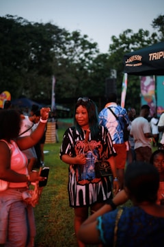 A group of people gathered outdoors during a festival or event. Vendors and individuals are interacting, some holding drinks or having conversations. There are tents and banners in the background with trees surrounding the area. The scene suggests a relaxed and lively atmosphere.