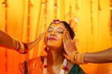 A woman with her eyes closed is having turmeric applied on her face by two people. She is adorned with floral jewelry and wears henna on her hands. The background is decorated with bright yellow and orange drapes, and she appears to be in a festive or ceremonial setting.