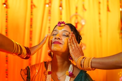 A woman with her eyes closed is having turmeric applied on her face by two people. She is adorned with floral jewelry and wears henna on her hands. The background is decorated with bright yellow and orange drapes, and she appears to be in a festive or ceremonial setting.