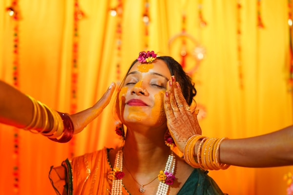 A woman with her eyes closed is having turmeric applied on her face by two people. She is adorned with floral jewelry and wears henna on her hands. The background is decorated with bright yellow and orange drapes, and she appears to be in a festive or ceremonial setting.