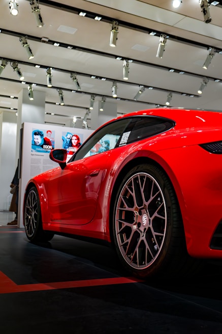 A sleek, red sports car is showcased indoors, surrounded by modern lighting fixtures on the ceiling. The focus is on the car's rear wheel and side, displaying intricate rim design. In the background, there are posters with images and descriptions of people, adding an element of exhibition or event atmosphere.