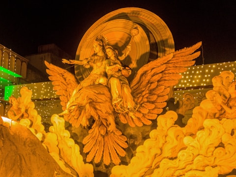 A detailed sculpture of two figures riding an enormous winged creature, set against an ornate backdrop. The figures are dramatically posed, with elaborate costumes and headdresses. The sculpture is brightly illuminated, highlighting its intricate details and the surrounding cloud-like formations. The background features a disk-like structure and fairy lights, enhancing the grandeur.