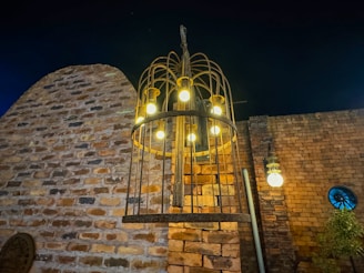 An outdoor setting with a large, rustic metal chandelier featuring multiple glowing light bulbs. The background consists of a weathered brick wall with varied hues, and there's a smaller light fixture on the wall. A tree or shrub is visible in the lower right corner, adjacent to a blue circular window.