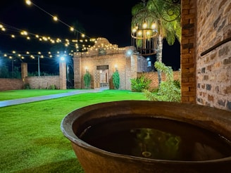 A night scene featuring a well-lit outdoor courtyard with a grassy lawn and a rustic brick structure in the background. String lights hang overhead, adding a warm glow to the setting. On the right is a large, round basin with water, and decorative plants are visible around the courtyard.