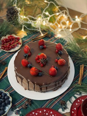 A richly decorated chocolate cake topped with fresh strawberries, blueberries, and red currants. The cake is set on a festive table surrounded by sprigs of pine, string lights, and additional bowls of berries. The ambiance is cozy and holiday-themed, with natural elements and warm lighting.