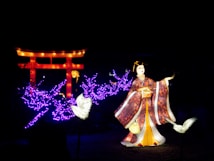 A brightly lit display featuring a traditional Japanese figure in a kimono surrounded by glowing purple lights that resemble tree branches. A large, illuminated red torii gate stands in the background. Two luminous white cranes are positioned near the figure, adding to the ethereal scene.