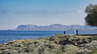 A scenic coastal view with a rocky shoreline covered in green shrubs. Two people are standing near a flagpole with flags, against a backdrop of blue sea and distant mountains under a clear sky.