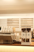 A modern coffee shop counter features a large metallic espresso machine with several stacked takeaway cups on top. Next to it, two white coffee grinders filled with beans are placed, labeled with origin signs depicting Colombia and Brazil. Above the counter, a menu board displays various drink options, including lemonade, chai latte, and hot chocolate.