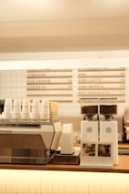 A modern coffee shop counter features a large metallic espresso machine with several stacked takeaway cups on top. Next to it, two white coffee grinders filled with beans are placed, labeled with origin signs depicting Colombia and Brazil. Above the counter, a menu board displays various drink options, including lemonade, chai latte, and hot chocolate.