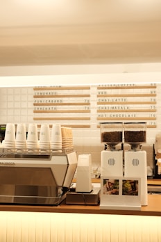 A modern coffee shop counter features a large metallic espresso machine with several stacked takeaway cups on top. Next to it, two white coffee grinders filled with beans are placed, labeled with origin signs depicting Colombia and Brazil. Above the counter, a menu board displays various drink options, including lemonade, chai latte, and hot chocolate.