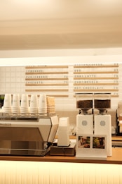 A modern coffee shop counter features a large metallic espresso machine with several stacked takeaway cups on top. Next to it, two white coffee grinders filled with beans are placed, labeled with origin signs depicting Colombia and Brazil. Above the counter, a menu board displays various drink options, including lemonade, chai latte, and hot chocolate.