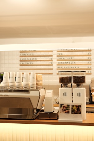 A modern coffee shop counter features a large metallic espresso machine with several stacked takeaway cups on top. Next to it, two white coffee grinders filled with beans are placed, labeled with origin signs depicting Colombia and Brazil. Above the counter, a menu board displays various drink options, including lemonade, chai latte, and hot chocolate.