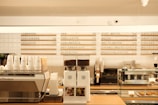 A modern coffee shop countertop with a variety of beverage options listed on the wall behind. The counter features a coffee machine, stacked paper cups, a coffee grinder with beans, and a display case with pastries. The overall aesthetic is clean and organized, with a neutral color palette.