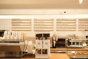A modern coffee shop countertop with a variety of beverage options listed on the wall behind. The counter features a coffee machine, stacked paper cups, a coffee grinder with beans, and a display case with pastries. The overall aesthetic is clean and organized, with a neutral color palette.