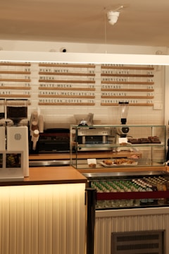 A café counter with various equipment such as a coffee grinder, espresso machine, and a display case containing pastries. Behind the counter, a menu board lists different types of coffee and their prices. The ambiance of the café is modern with warm lighting and a clean layout.