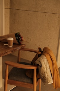 A cozy wooden chair with a gray cushion is placed next to a wooden table. On the table, there is a paper-wrapped bouquet of flowers and a takeaway coffee cup. A coat and a knitted bag hang over the backrest of the chair, lending a warm and inviting feel.