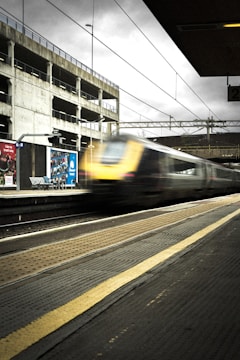 A train station platform with a high-speed train passing by in a blur. The station is a mix of concrete and metal structures, with overhead electrical lines visible. There is a parking structure in the background, and overcast skies add to the atmosphere. The platform has tactile paving and signage indicating platform numbers.