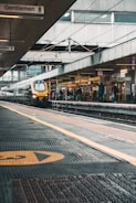 A busy train station platform with a train moving quickly in the foreground. The station features overhead signs indicating directions, with several passengers waiting on the platform. The platform is covered, with visible structures and beams, contributing to an urban transportation setting.
