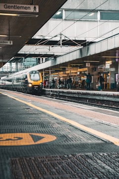 A busy train station platform with a train moving quickly in the foreground. The station features overhead signs indicating directions, with several passengers waiting on the platform. The platform is covered, with visible structures and beams, contributing to an urban transportation setting.