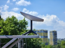 A satellite dish is mounted on a metal structure with a background of green trees and modern high-rise buildings. The sky is clear with scattered clouds, suggesting a pleasant weather.