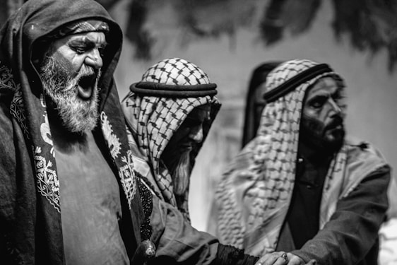 Three men are depicted wearing traditional Middle Eastern attire, including keffiyehs and robes. One man is visibly expressive, possibly shouting or singing, while the other two appear more subdued or introspective. The image is in black and white, adding a dramatic and timeless feel to the scene.