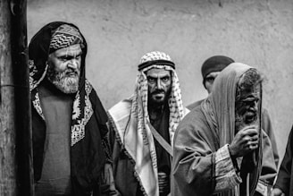 Four men in traditional Middle Eastern attire are gathered, each displaying serious or intense expressions. The clothing includes headscarves and robes, and the scene is captured in black and white, emphasizing the textures and contrasts.