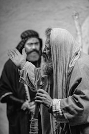A black and white image depicts two people wearing robes that suggest historical or biblical attire. The person in the foreground, with a beard, is gesturing with one hand while holding a staff with the other. Another person is visible in the background, slightly out of focus, wearing a headdress.