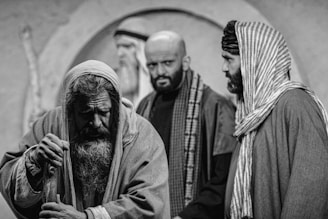 Three men are depicted in a dramatic black and white setting, dressed in historical or biblical clothing. The man in the foreground has a long beard and appears weary or introspective, leaning on a staff. The two men in the background are looking in different directions, with expressions that convey seriousness or contemplation.