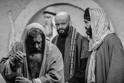 Three men are depicted in a dramatic black and white setting, dressed in historical or biblical clothing. The man in the foreground has a long beard and appears weary or introspective, leaning on a staff. The two men in the background are looking in different directions, with expressions that convey seriousness or contemplation.