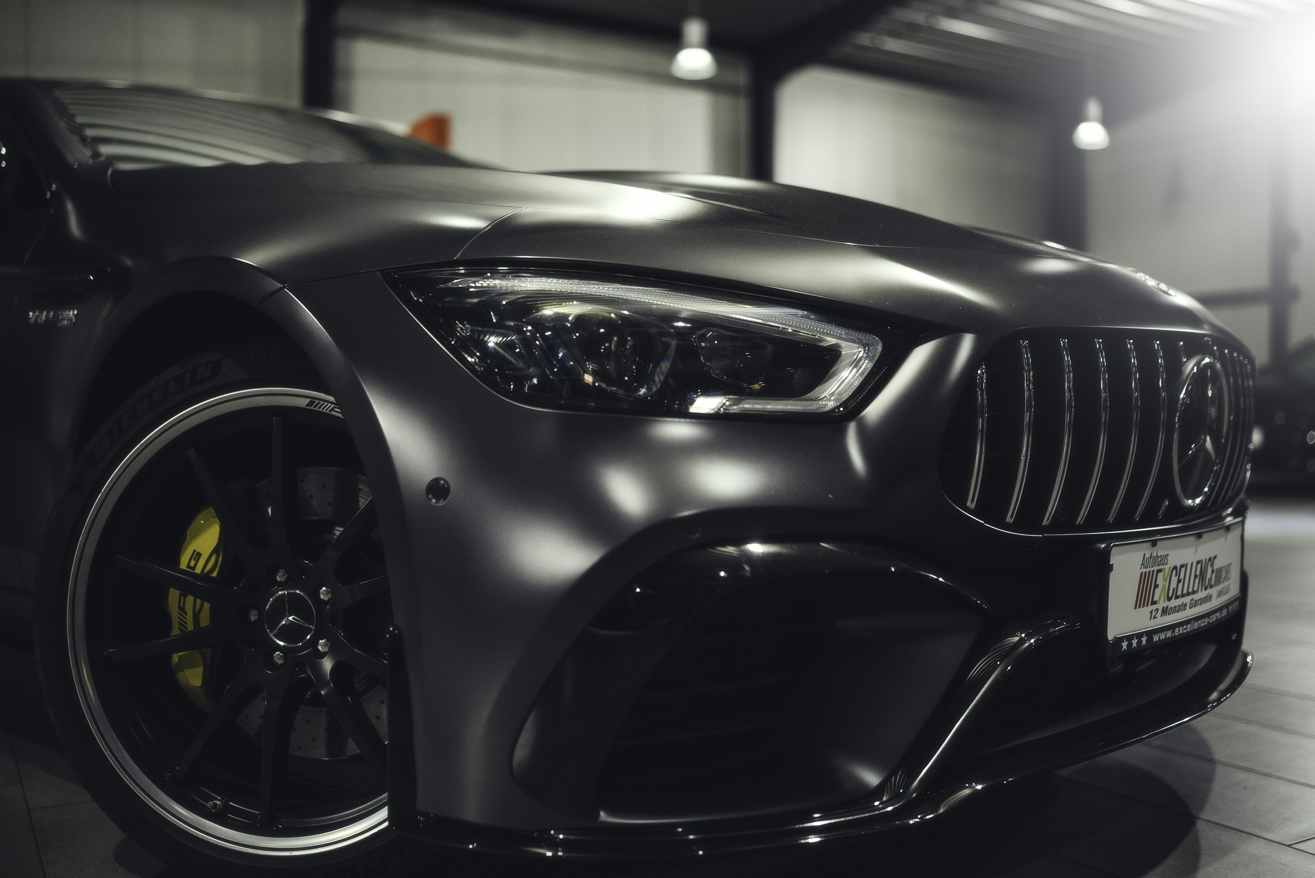 A close-up view of the front part of a sleek, modern car with a metallic finish, showcasing the intricate details of the front headlight, grille, and the emblem on the rim. The car is parked indoors under soft lighting, highlighting its glossy surface and design features.