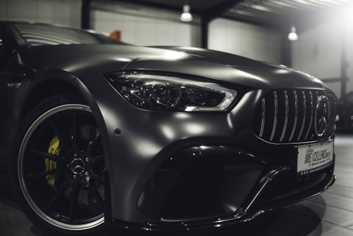 A close-up view of the front part of a sleek, modern car with a metallic finish, showcasing the intricate details of the front headlight, grille, and the emblem on the rim. The car is parked indoors under soft lighting, highlighting its glossy surface and design features.