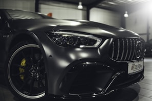 A close-up view of the front part of a sleek, modern car with a metallic finish, showcasing the intricate details of the front headlight, grille, and the emblem on the rim. The car is parked indoors under soft lighting, highlighting its glossy surface and design features.