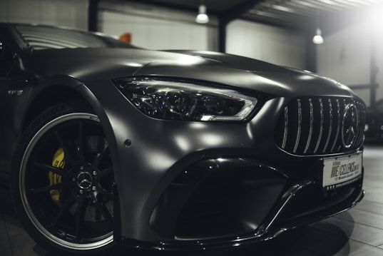 A close-up view of the front part of a sleek, modern car with a metallic finish, showcasing the intricate details of the front headlight, grille, and the emblem on the rim. The car is parked indoors under soft lighting, highlighting its glossy surface and design features.