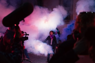 A street scene filled with dense smoke, illuminated by colorful lighting, with a central focus on a person standing amid the fog. Around, people are holding cameras and a microphone setup, suggesting a possible filming or protest event.