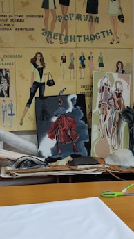 The image portrays a fashion design workspace with illustrated fashion sketches on a wall featuring various outfits. There are two canvases with vibrant artwork, one depicting a figure in a red dress. The surface is cluttered with papers, fabric rolls, and cutting tools.