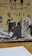 The image portrays a fashion design workspace with illustrated fashion sketches on a wall featuring various outfits. There are two canvases with vibrant artwork, one depicting a figure in a red dress. The surface is cluttered with papers, fabric rolls, and cutting tools.