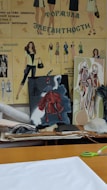The image portrays a fashion design workspace with illustrated fashion sketches on a wall featuring various outfits. There are two canvases with vibrant artwork, one depicting a figure in a red dress. The surface is cluttered with papers, fabric rolls, and cutting tools.