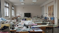 A well-lit room filled with sewing machines and various sewing materials scattered across large tables. The walls have sketches of fashion designs, and there's a chalkboard at the far end. The windows let in natural light, illuminating the workspace.