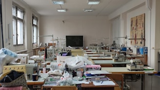 A well-lit room filled with sewing machines and various sewing materials scattered across large tables. The walls have sketches of fashion designs, and there's a chalkboard at the far end. The windows let in natural light, illuminating the workspace.