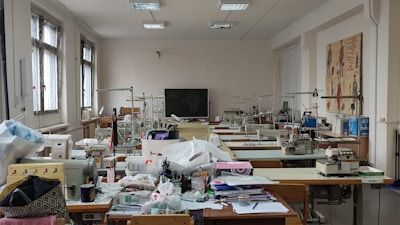 A well-lit room filled with sewing machines and various sewing materials scattered across large tables. The walls have sketches of fashion designs, and there's a chalkboard at the far end. The windows let in natural light, illuminating the workspace.