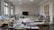 A well-lit room filled with sewing machines and various sewing materials scattered across large tables. The walls have sketches of fashion designs, and there's a chalkboard at the far end. The windows let in natural light, illuminating the workspace.