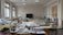 A well-lit room filled with sewing machines and various sewing materials scattered across large tables. The walls have sketches of fashion designs, and there's a chalkboard at the far end. The windows let in natural light, illuminating the workspace.