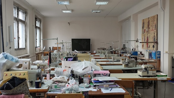 A well-lit room filled with sewing machines and various sewing materials scattered across large tables. The walls have sketches of fashion designs, and there's a chalkboard at the far end. The windows let in natural light, illuminating the workspace.