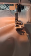 A sewing machine needle is stitching fabric. The machine is partially visible with threads attached and the fabric is smooth and light-colored, being guided through the machine's foot.