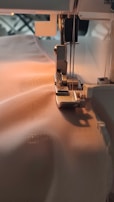 A sewing machine needle is stitching fabric. The machine is partially visible with threads attached and the fabric is smooth and light-colored, being guided through the machine's foot.