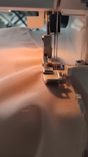 A sewing machine needle is stitching fabric. The machine is partially visible with threads attached and the fabric is smooth and light-colored, being guided through the machine's foot.