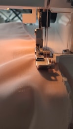 A sewing machine needle is stitching fabric. The machine is partially visible with threads attached and the fabric is smooth and light-colored, being guided through the machine's foot.