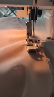A sewing machine needle is stitching fabric. The machine is partially visible with threads attached and the fabric is smooth and light-colored, being guided through the machine's foot.