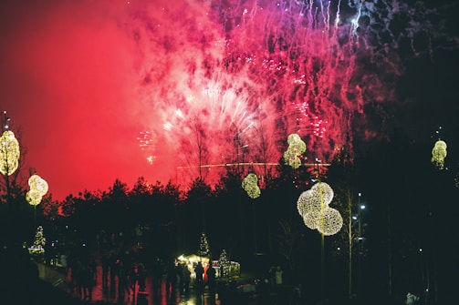 Vibrant fireworks burst in the night sky, casting a red glow over a public park. Illuminated decorations hang from the trees, creating a festive atmosphere. A crowd of people gathers below, watching the display while surrounded by glowing lights.