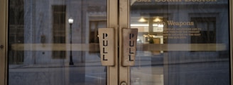 Two glass doors with large pull handles, framed by an exterior urban setting. Visible text on one door includes a 'No Weapons' policy and instructions for deliveries.